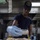 170123-N-PP996-072 A person wearing a dark navy cap with an insignia and blue latex gloves is seasoning a large tray of raw chicken pieces in what appears to be a commercial or military galley kitchen. They are holding a spice/seasoning shaker and applying it to the chicken, which is already visibly coated with herbs and spices. The industrial kitchen environment and military-style cap suggest this may be a naval vessel or military food service setting.