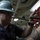 170118-N-BR087-001 A U.S. Navy sailor wearing a white hard hat marked '74' and safety glasses is performing maintenance work in what appears to be a ship's engineering or mechanical space. He is using a red pipe wrench or similar tool, working alongside another person whose tattooed arm is partially visible. The setting appears to be an interior mechanical compartment of a naval vessel.