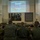 HMH-464 Marines attend Road to War Brief during Exercise Frigid Condor A military briefing or training session is taking place in what appears to be a military facility. Two uniformed personnel stand at the front presenting a projected slideshow to a group of seated military members. The presentation slide displays 'Questions?' and references a fictional country 'Bantutkistan' marked as 'Overall Classification: UNCLASSIFIED'.