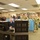170123-N-TI693-123 A military operations or command center facility showing a group of U.S. Navy personnel in both blue service uniforms and digital camouflage (NWU) uniforms gathered around computer workstations. Several personnel appear to be observing something off-camera, possibly a briefing or demonstration. The setting appears to be a training or command facility with multiple computer terminals and blue equipment racks.