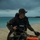 31st Marine Expeditionary Unit completes MEUEX A military or special operations personnel is kneeling on a sandy beach after an apparent amphibious insertion. The individual is equipped with camouflage uniform, tactical vest, weapon, and diving/swimming gear including fins and what appears to be an inflatable flotation device. The scene suggests a combat swimmer or reconnaissance operation with overcast skies and turquoise water in the background.