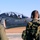 Every clime and place: VMAT-203 Marines maintain expeditionary mindset at MCALF Bogue Two military personnel in flight suits and camouflage uniforms observe a military jet aircraft taxiing on a runway. The aircraft appears to be a two-seat trainer or attack jet with two occupants visible in the cockpit wearing flight helmets. The scene takes place on a military airfield during daylight hours under clear blue skies.