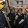 170117-N-OA516-0006 A group of naval personnel in firefighting gear are gathered in what appears to be a ship's interior compartment, likely during a damage control drill or training exercise. They are wearing red hard hats with face shields, flash hoods, navy blue coveralls, and protective gloves. The setting shows typical shipboard piping, valves, and structural elements in the background.