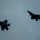 Lightning II strikes Iwakuni, F-35B arrives Two fifth-generation stealth fighter jets, identifiable as F-35 Lightning II aircraft, are photographed in flight against an overcast gray sky. The aircraft appear in near-silhouette due to low-light conditions, with one aircraft in the foreground-left and one in the background-right. Faint vapor or contrail lines are visible trailing from the aircraft.