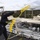 170123-N-KP948-104 A sailor aboard a naval vessel is throwing a heaving line with a red monkey fist knot weighted ball toward a pier or dock. The yellow line is mid-flight, captured in motion against a backdrop of a naval fuel station facility. A second uniformed personnel is visible walking on the pier below.