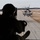 HMH-464 Marines Conduct Flight Operations during Exercise Frigid Condor A silhouetted military crew member is seated at the open rear ramp of a helicopter, observing or operating equipment while a large military helicopter marked '59' hovers low over a runway ahead. The scene appears to be a formation flight or tactical exercise conducted in cold-weather conditions, evidenced by patches of snow on the surrounding terrain. The photograph is taken from inside one aircraft looking out toward the second helicopter.