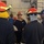 170117-N-OA516-0042 A group of military personnel, likely U.S. Navy sailors, are gathered in what appears to be a ship's interior compartment during a damage control or firefighting drill. Several individuals are wearing red firefighting helmets with yellow reflective strips and protective gear, while at least one person in a dark uniform with a U.S. flag patch appears to be an instructor or supervisor. A central figure in firefighting gear appears to be speaking or receiving instruction.
