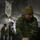 2d Medical Bn Field Exercise A military medical corpsman in camouflage uniform and olive beanie is treating a casualty on a field stretcher inside a military medical tent. The patient appears to have visible wounds and is receiving IV fluid treatment along with wound dressing. Additional military personnel are visible in the background, suggesting a field hospital or combat casualty care exercise.