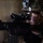 2/5 Mechanized Raid A military service member in MARPAT (Marine pattern) camouflage uniform is aiming down the optic sight of an assault rifle, likely an M4 carbine or M16 variant. The individual is wearing a combat helmet with a night vision device mount and has tactical face paint applied. The setting appears to be an indoor or low-light environment against a concrete or adobe wall.
