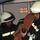161229-N-N0901-757 Two firefighters in full turnout gear with white helmets and high-visibility reflective striping are handling a folded fire hose with a coupling/nozzle end. They appear to be in a fire station or apparatus bay, with a digital display board visible in the background showing 'FPL 4-51' and 'COV 4-49'. The scene suggests equipment preparation, inspection, or deployment activity.