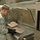 A U.S. Air Force airman in ABU (Airman Battle Uniform) camouflage is working in a military dining facility kitchen. The airman is handling what appear to be food trays or packaged meal items on a large commercial steam cooking appliance. Steam is visibly rising from the equipment, indicating active cooking operations.