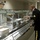 A military dining facility (DFAC/mess hall) scene showing two service members interacting at a food service line. A junior enlisted Airman in ABU (Airmen Battle Uniform) with blue food-service gloves is serving food behind the counter, while a U.S. Army Sergeant in Army Service Uniform (ASU, dress blacks) is receiving food on a tray. The setting appears to be a modern military installation cafeteria.