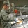 A U.S. Air Force airman in ABU (Airman Battle Uniform) is serving food at a military dining facility (DFAC). The airman is wearing blue food-service gloves and handling a food tray at a stainless steel serving line. The setting appears to be a standard military chow hall with commercial-grade food service equipment.