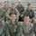 A group of nine U.S. Air Force personnel posing for a group photo on bleachers, wearing ABU (Airman Battle Uniform) camouflage and gray fleece jackets. Name tapes are visible on several individuals, including Thomas, Sagstetter, Shannon, Jacobson, Welsh, and Timberman. The setting appears to be an indoor gymnasium or sports facility.
