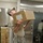 A military service member in US Air Force Airman Battle Uniform (ABU) is lifting a large cardboard box overhead in what appears to be a military dining facility or supply area. A second uniformed individual is visible in the background near what appears to be a large stainless steel oven or equipment unit. Multiple stacked cardboard boxes on a hand truck are visible to the left.