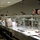 The image shows a military dining facility (DFAC/mess hall) with service members in camouflage uniforms going through a food service line. A uniformed server wearing blue gloves is working behind the counter on the left side. The stainless steel serving line contains food trays, fruit, and other items visible under sneeze guards.