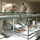 Two U.S. Air Force personnel in Airman Battle Uniform (ABU) camouflage pattern are serving food at a military dining facility (DFAC/chow hall). They are working behind a standard institutional cafeteria-style serving line with a sneeze guard. A third individual in a white cook's uniform is visible in the background through a service window.