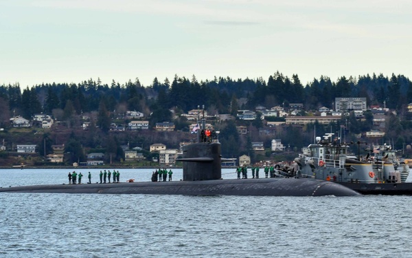 A large military submarine is transiting on calm waters, accompanied by a naval tugboat on its starboard side. Crew members dressed in green uniforms are visible standing on the submarine's deck, suggesting a port arrival or departure evolution. A residential shoreline with trees and buildings is visible in the background.