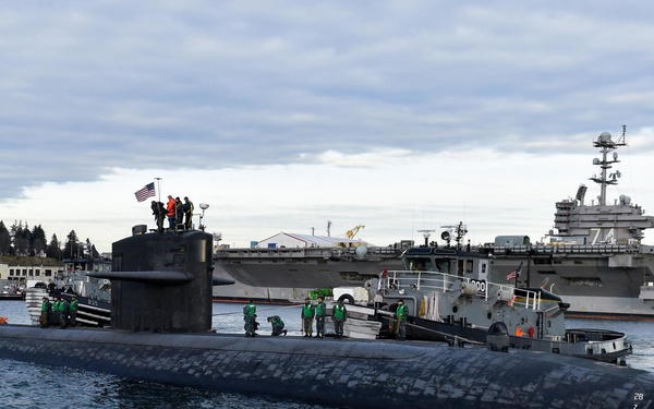 A U.S. Navy submarine is docking or maneuvering at a naval base, assisted by a tugboat alongside its hull. Crew members in green vests are stationed along the submarine's deck, with personnel also visible on the sail (conning tower) flying an American flag. In the background, a large aircraft carrier bearing the number '74' is moored at the pier.