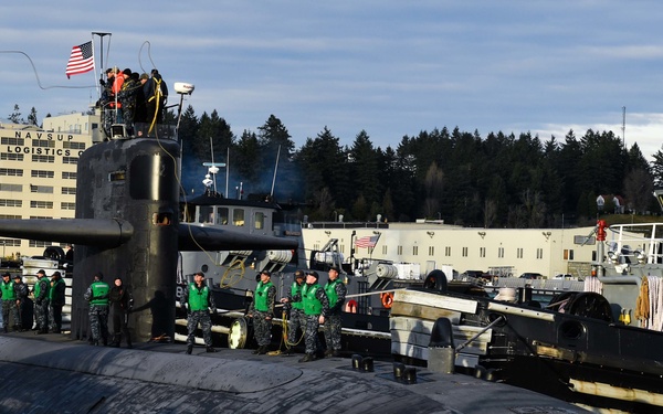 A U.S. Navy submarine is docked at a naval installation, identifiable by the 'NAVSUP Fleet Logistics' building visible in the background. Numerous sailors in green vests and Navy working uniforms are stationed along the hull of the submarine, suggesting a homecoming, departure, or formal docking evolution. A support tugboat numbered '800' is positioned alongside the submarine on the starboard side.