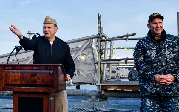 Two U.S. Navy personnel are positioned on what appears to be the deck of a submarine or naval vessel. One officer in a combination cover and khaki trousers stands at a wooden podium speaking into a microphone, while a second sailor in Navy Working Uniform (NWU) Type I digital camouflage stands at ease nearby. The setting appears to be an outdoor ceremony or briefing at a naval facility.