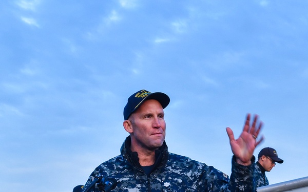 A U.S. Navy officer in Navy Working Uniform (NWU Type I digital camouflage) stands at a wooden podium and addresses an audience, with one hand raised in a gesturing motion. The setting appears to be an outdoor naval facility or ship deck, likely during an official ceremony or briefing. A second uniformed Navy personnel member is visible in the background near what appears to be a gangway or brow structure.