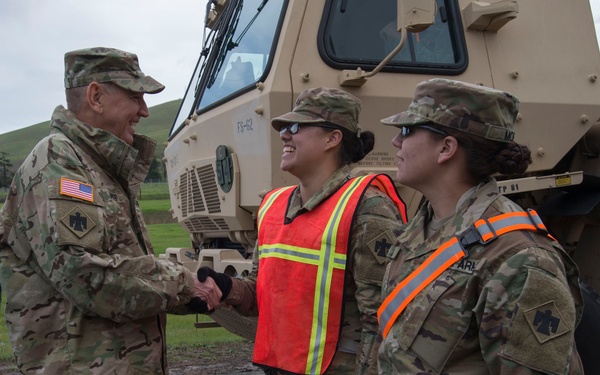 A senior U.S. Army officer is shaking hands with a female soldier wearing a high-visibility orange safety vest, while a second female soldier stands nearby. All three individuals are wearing MultiCam (OCP) camouflage uniforms and patrol caps, with a tan military vehicle visible in the background. The interaction appears to be a formal greeting or commendation during a field exercise or unit inspection.