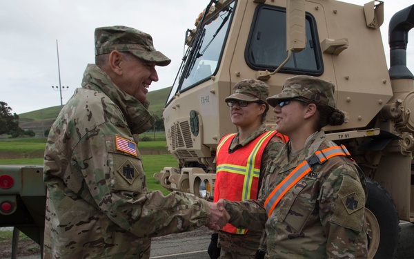 A senior U.S. Army officer in Operational Camouflage Pattern (OCP) uniform is shaking hands with a soldier wearing a high-visibility safety vest, while a second soldier stands nearby. All personnel wear OCP uniforms and display a unit patch on their left shoulder. A large tan military tactical vehicle is visible in the background.