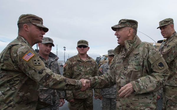 A group of U.S. Army soldiers in MultiCam/OCP camouflage uniforms are gathered in an outdoor parking area. Two soldiers in the foreground are shaking hands, suggesting a greeting, farewell, or recognition ceremony. Military vehicles are visible in the background under an overcast sky.