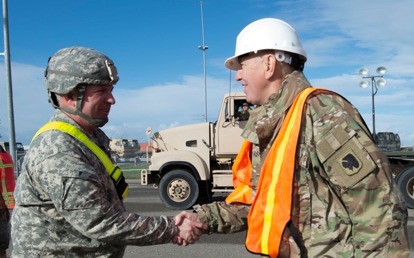 Two military personnel in uniform are shaking hands in what appears to be a military staging or port operations area. The soldier on the left wears ACU (Army Combat Uniform) with a combat helmet and yellow safety vest, while the soldier on the right wears OCP (Operational Camouflage Pattern) uniform with an orange safety vest and white hard hat. Military trucks and vehicles are visible in the background.