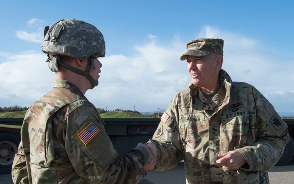 Two U.S. Army soldiers in MultiCam/OCP camouflage uniforms are shaking hands outdoors on a paved surface. The soldier on the right, identified by a name tape reading 'ASHER' and wearing a patrol cap, appears to be a senior officer. The soldier on the left is wearing a combat helmet and has a U.S. flag patch on the right sleeve.