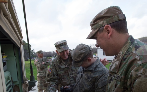 Several U.S. Army soldiers in OCP (Operational Camouflage Pattern) uniforms are gathered around a military vehicle, working on or inspecting equipment on a flat surface. The scene appears to be a maintenance or training exercise conducted outdoors at what looks like a military installation. At least one soldier is smiling, suggesting a collaborative and relatively relaxed working environment.