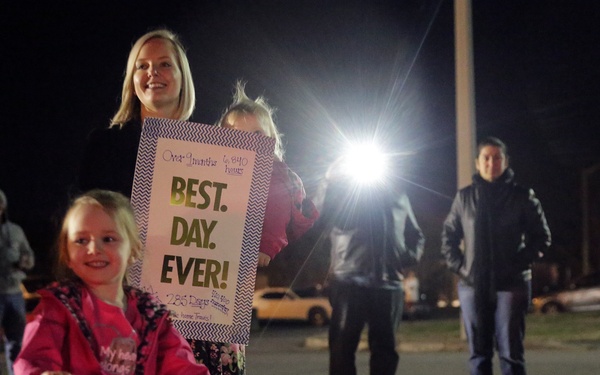 A nighttime homecoming scene showing a woman and young girl holding handmade signs welcoming a military or deployed family member home. The young girl holds a bright pink sign reading 'WELCOME HOME DADDY' while the woman behind her holds a sign reading 'BEST. DAY. EVER.' Several other people stand in the background near a parking area illuminated by bright lights.