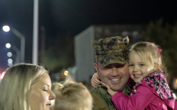 A U.S. Marine in digital camouflage uniform and cover (cap) is reuniting with his family at night, likely following a deployment. He is holding a young girl in a pink jacket while a woman, presumably his wife, holds a toddler nearby. The scene appears to be a military homecoming event on a base, with other people and vehicles visible in the background.