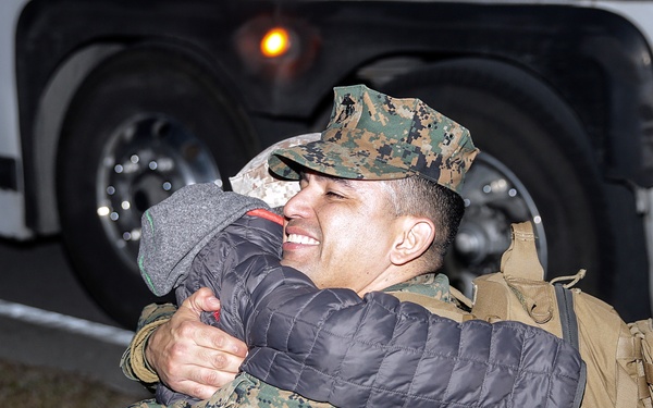 A U.S. Marine in woodland MARPAT camouflage uniform and cover is embracing a civilian in a dark quilted jacket, both appearing to share an emotional reunion. The Marine is smiling broadly and carrying a tan tactical backpack. The scene appears to take place at night near a large white vehicle, likely a bus.
