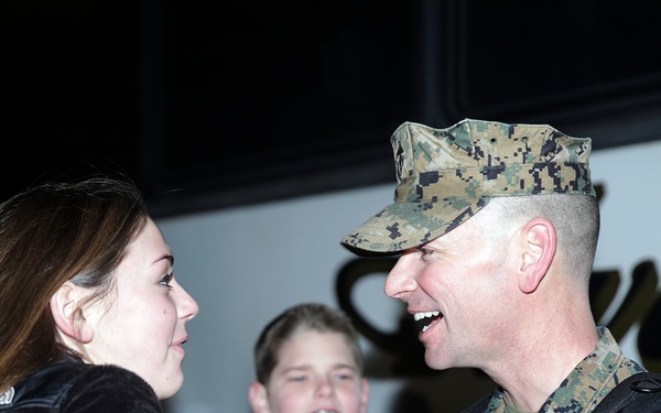 A U.S. Marine in woodland MARPAT camouflage uniform and cover is smiling broadly while greeting a young woman who has her fist raised toward his chest, suggesting a playful or affectionate interaction. A young boy is visible in the background, also smiling. The setting appears to be near a bus or large vehicle, consistent with a military homecoming or departure event.