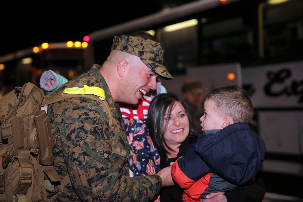 A U.S. Marine in digital camouflage uniform is reuniting with a woman and a young child at night, likely following a deployment return. The Marine is smiling and leaning toward the child, who is being held by the woman. A large bus is visible in the background, consistent with a military homecoming event.