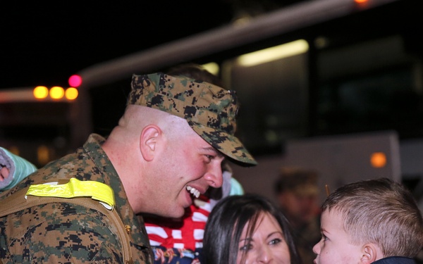 A U.S. Marine in digital camouflage uniform is reuniting with a woman and a young child at night, likely following a deployment return. The Marine is smiling and leaning toward the child, who is being held by the woman. A large bus is visible in the background, consistent with a military homecoming event.