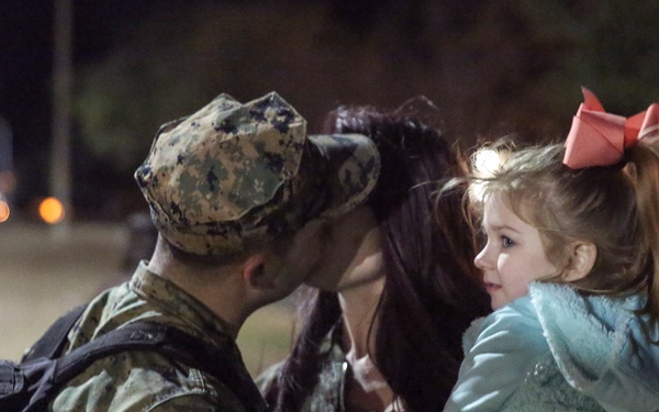 A military service member in camouflage uniform shares an emotional moment with a woman and a young child at night. The scene appears to depict a farewell or homecoming, with the service member leaning in close to the child and woman. The nighttime outdoor setting with bokeh lights in the background adds an intimate atmosphere to the moment.