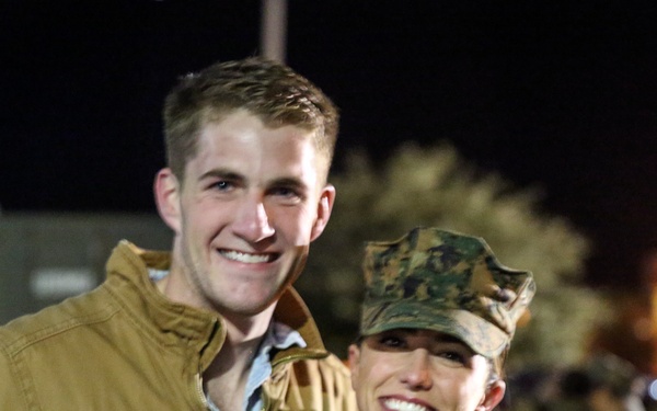 Two people pose together smiling at night in an outdoor setting. One individual is wearing U.S. Marine Corps MARPAT (woodland digital camouflage) uniform and cover (cap), while the other is in civilian clothing. The background shows a crowd of people and artificial lighting consistent with a military base or event.