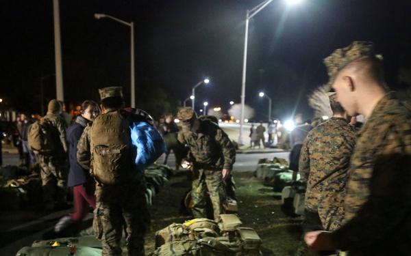 A nighttime military assembly showing personnel in camouflage uniforms gathered around equipment and gear laid out on the ground. The scene appears to be a staging or inspection area illuminated by street lights. Multiple military personnel are moving among rows of tactical backpacks, duffel bags, and field equipment.