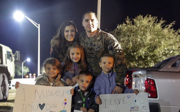 A military serviceman in camouflage uniform is reunited with his family at night, likely on a military base. His wife and four young children are holding handmade signs welcoming him home. The emotional scene captures a homecoming moment with signs reading 'I Missed You Dad,' 'Best Day Ever,' and 'I Love You Dad.'