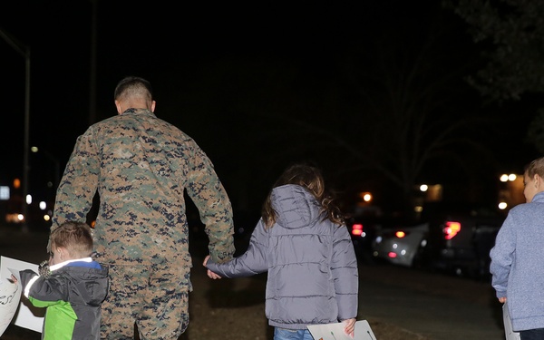 A military service member in camouflage uniform walks with several children at night, holding hands with them. The children are carrying handmade welcome signs, suggesting this is a homecoming or reunion event. The scene takes place outdoors in a parking or gathering area with street lights visible in the background.