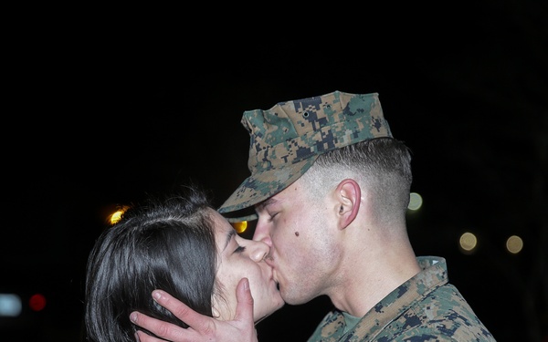 A person in U.S. Marine Corps MARPAT digital camouflage uniform and cover (cap) shares a kiss with a civilian at night. The setting appears to be an outdoor area, possibly a base or parking lot, with vehicle lights visible in the background. The scene is consistent with a military homecoming or farewell moment.