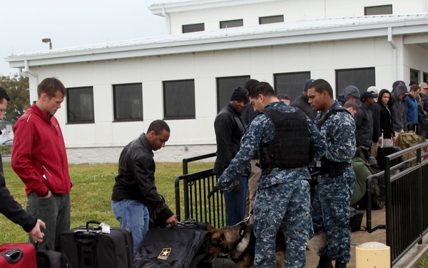 Military or security personnel in Navy digital camouflage uniforms are conducting a K-9 bag inspection at what appears to be a military installation entrance. A German Shepherd detection dog is actively sniffing luggage belonging to a line of civilians waiting to enter. The scene takes place outdoors on a wet, overcast day near a white institutional building.