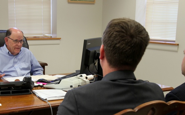 Three men are engaged in a meeting inside a traditional office setting. An older man seated behind a large wooden desk appears to be speaking or presenting to two younger men seated across from him. The setting suggests a formal business or professional consultation.