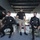 Army Divers plunge into the Pacific during exercise "Deep Blue" The image shows what appears to be a military or naval diving preparation scene aboard a vessel or facility. Two individuals in black wetsuits with diving harnesses are seated on benches, while a standing supervisor in a black t-shirt and khaki shorts oversees the preparation. A fourth person in the background appears to be assisting with equipment.