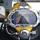 Army Divers plunge into the Pacific during exercise "Deep Blue" A person is wearing a professional Kirby Morgan diving helmet (model 37) with a yellow fiberglass shell and large chrome-framed viewport. The helmet is equipped with a Diving Systems International (DSI) regulator and various valves and fittings. The setting appears to be on a vessel or dock, with an EXIT sign visible in the background.