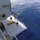 Army Divers plunge into the Pacific during exercise "Deep Blue" An aerial view taken from an elevated position on a large vessel shows three crew members standing on a lower deck platform extending over open ocean. A thick rope or cable extends from the ship diagonally into the deep blue water. The crew members appear to be managing lines or conducting deck operations at sea.