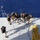 Army Divers plunge into the Pacific during exercise "Deep Blue" An aerial/overhead view of a ship's deck showing a group of personnel in matching dark shirts with yellow insignia preparing diving or underwater equipment near the edge of the vessel. The team appears to be conducting a coordinated operation involving ropes, lines, and what appear to be diving helmets or underwater equipment. The deep blue open ocean is visible beyond the deck edge.